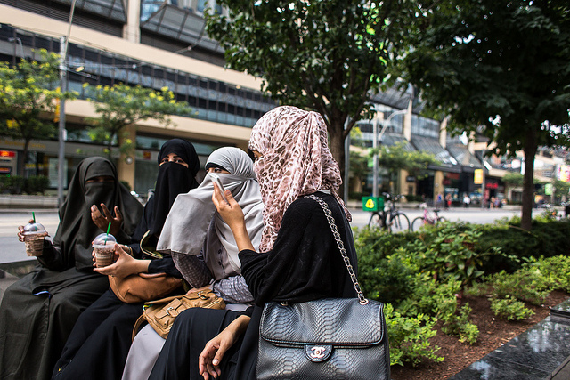 Four woman sit wearing hijabs on a planter near Bay and Dundas (August 2013).