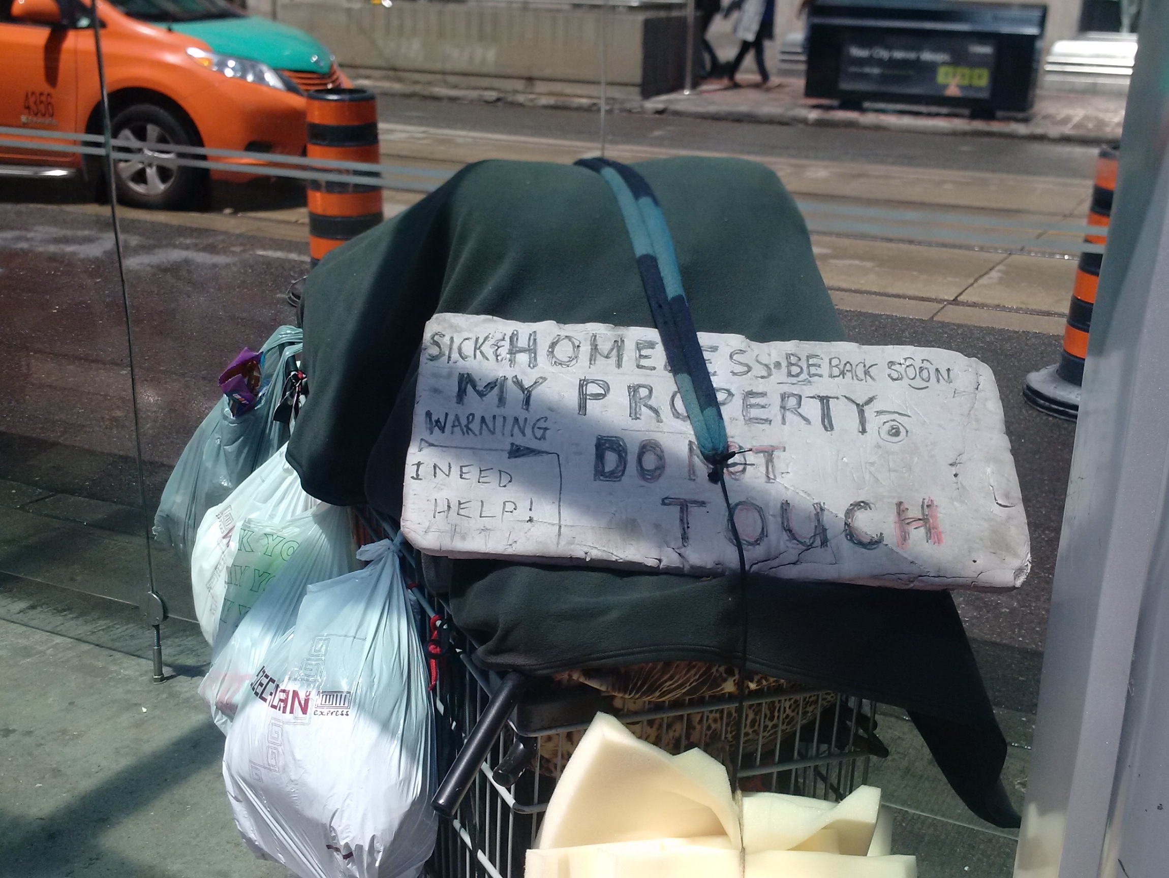 A homeless person's cart of belongings in a bus shelter has a sign on it that says "Sick and homeless. I need help."