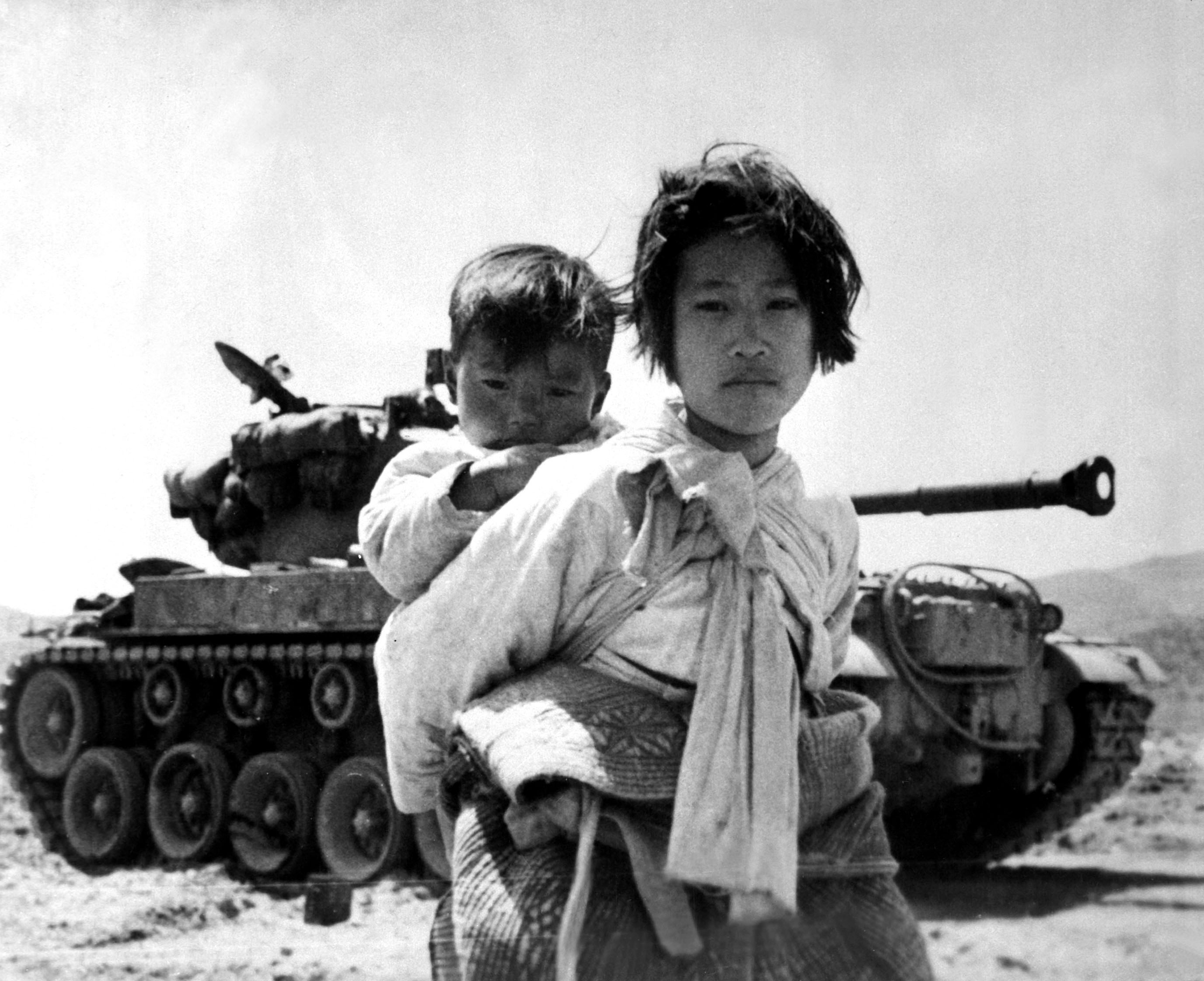 With her brother on her back a war weary Korean girl tiredly trudges by a stalled M-26 tank, at Haengju, Korea, in June 1951.