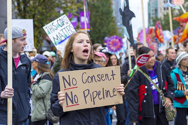 A protester holds a sign at a 2017 anti-pipeline rally in Vancouver. Image: William Chen/Wikimedia Commons