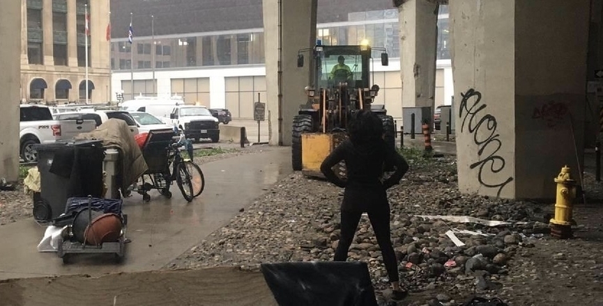 A teacher stands in front of a bulldozer at an encampment eviction under the Gardiner Expressway. Image: submitted by Cathy Crowe