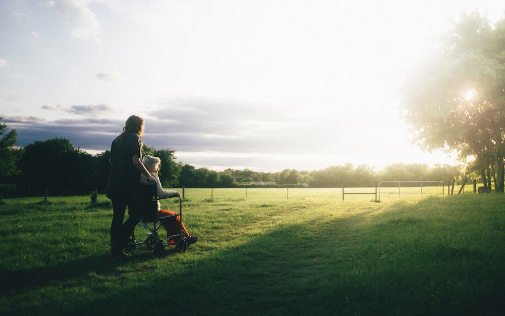 Person stands next to elderly person in a wheelchair outside in a field. Image credit: Dominik Lange/Unsplash