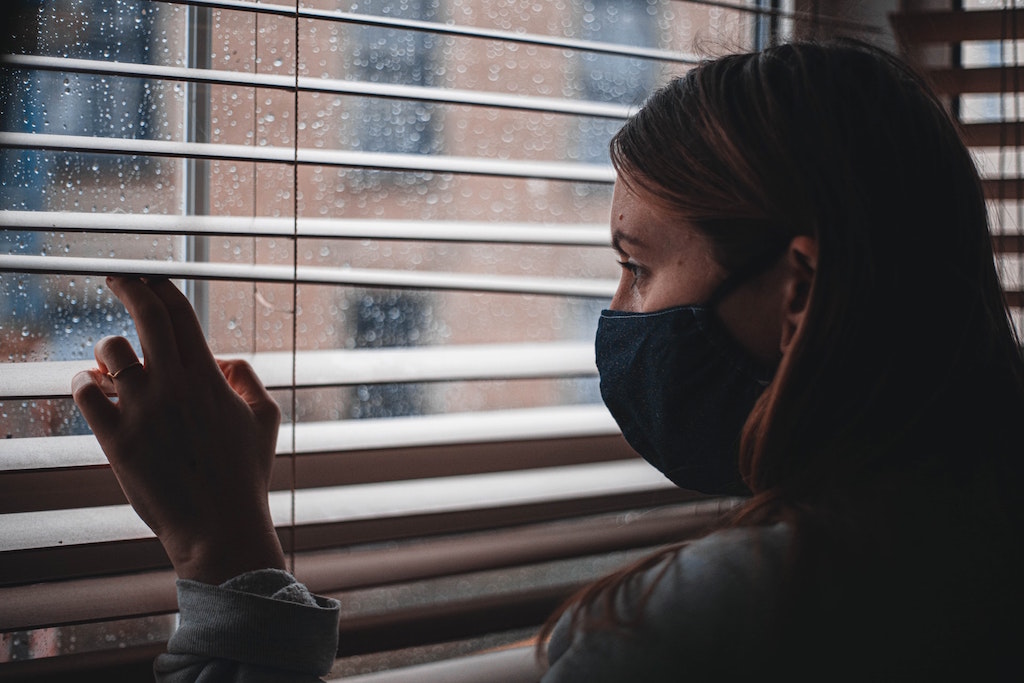 Person wearing mask looks out window. Image credit: Hedgehog Digital/Unsplash