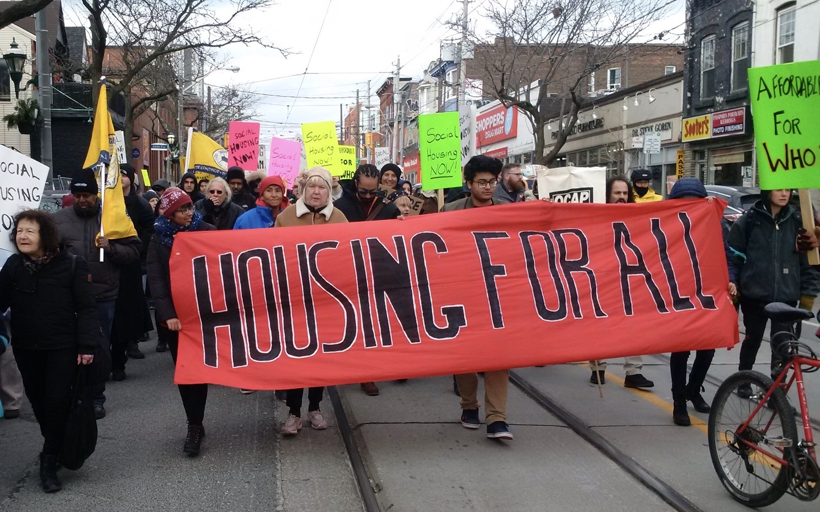 Housing advocates carry a banner. Black words on red read, "Housing for All." Many signs in background say "Social Housing Now." Image: Cathy Crowe/Used with permission