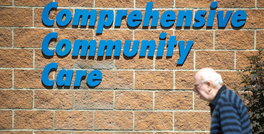 A sign reading "comprehensive community care" with an elderly man walking in front of it. Image: Government of Alberta/Flickr