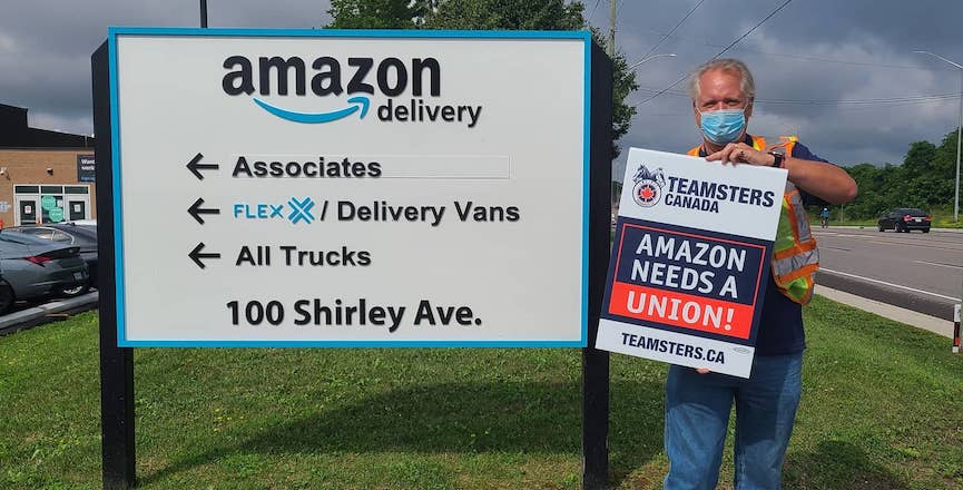 A member of Teamsters Local 879 stands in front of an Amazon warehouse sign holding a Teamsters Canada sign that reads "Amazon needs a union." Image: Teamsters Canada/Twitter