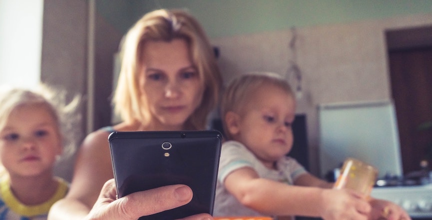 A woman holding a baby with a toddler on the other side focuses on her phone. Image: Vitolda Klein/Unsplash