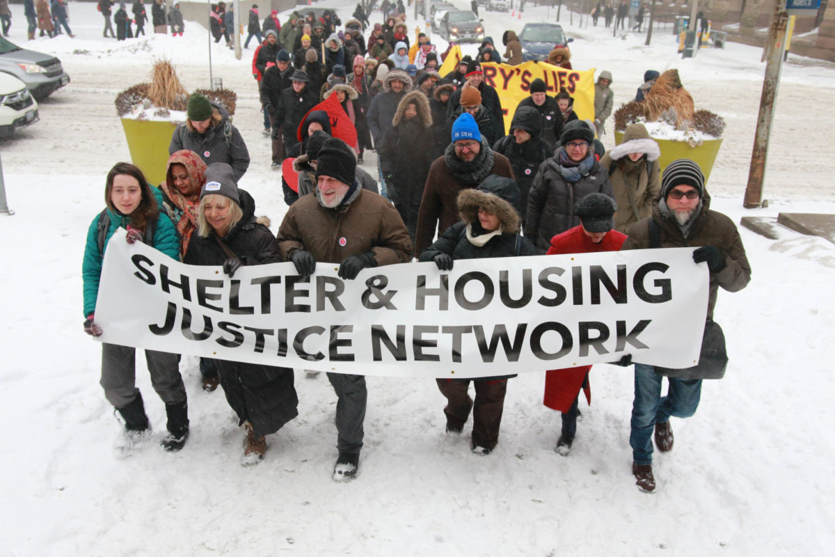 A group of people march behind the Shelter & Housing Justice Network banner in the snow. (Image: Paul Salvatori/Used with permission)