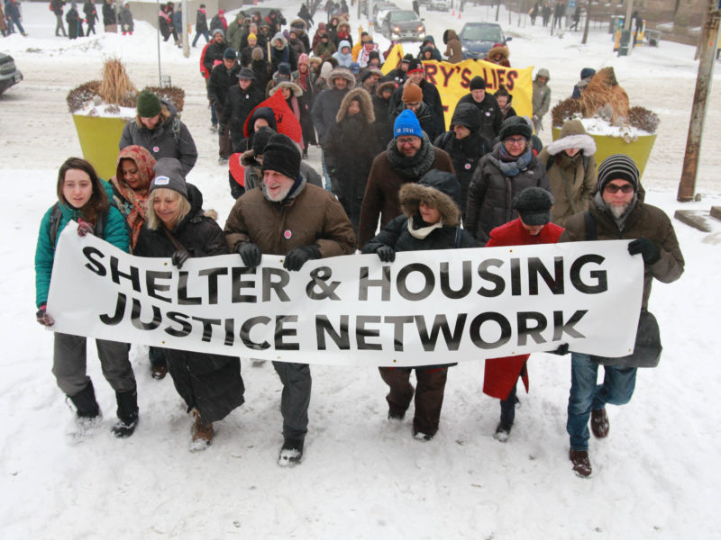 A group of people march behind the Shelter & Housing Justice Network banner in the snow. (Image: Paul Salvatori/Used with permission)