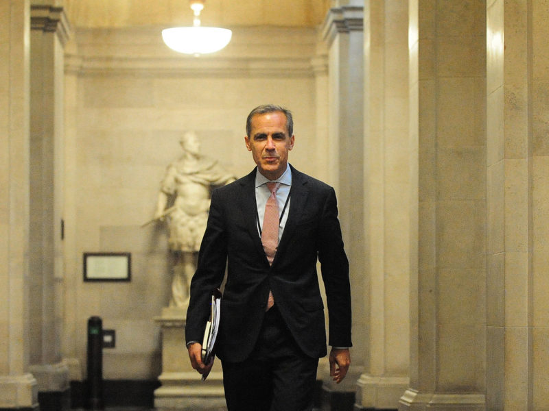 Mark Carney walks down a marble hallway with files under his arm.