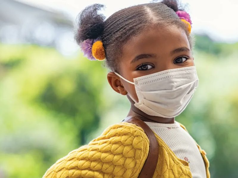 Image of a Black child with a yellow jacket wearing a medical mask outside.