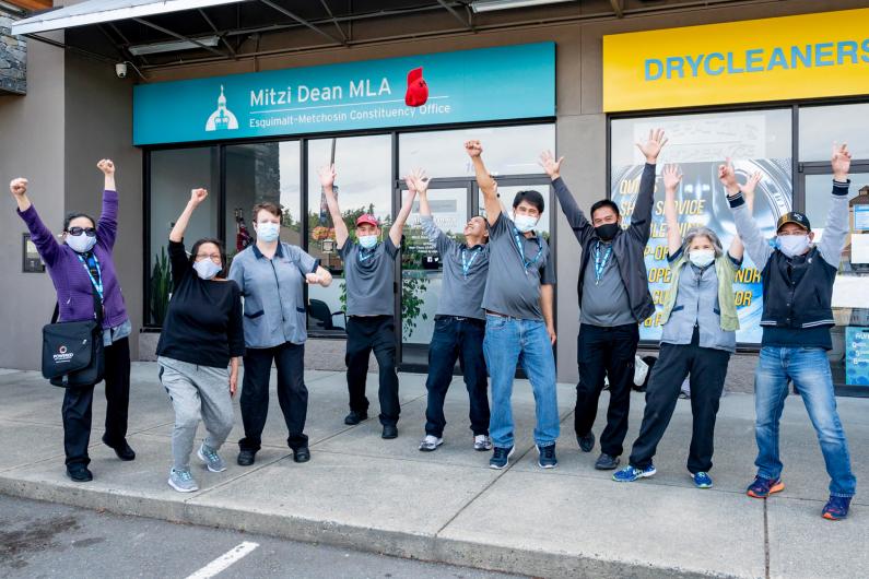 Photo of a group of people wearing medical masks and raising their fists to the sky in from of MLA Mitzi Dean's constituency office