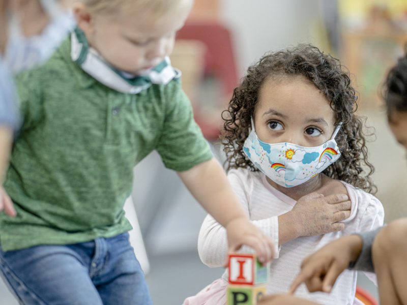 Group of mixed race children playing on the carpet at daycare while wearing reusable face coverings due to the new COVID-19 regulations.