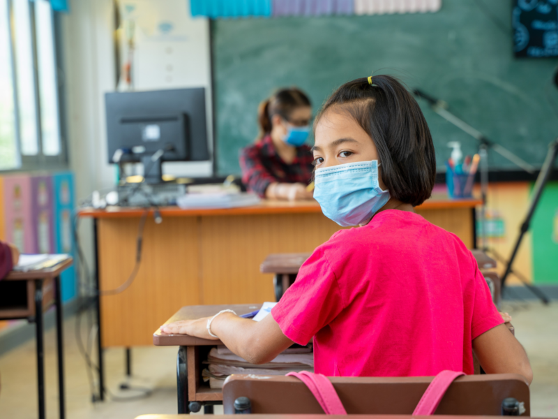 Photo of a young girl sitting in a classroom wearing a medical mask.