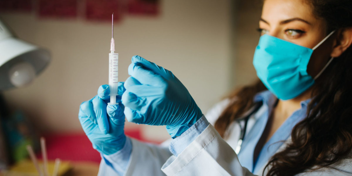 Health care worker, wearing a mask and gloves, holding a needle up.