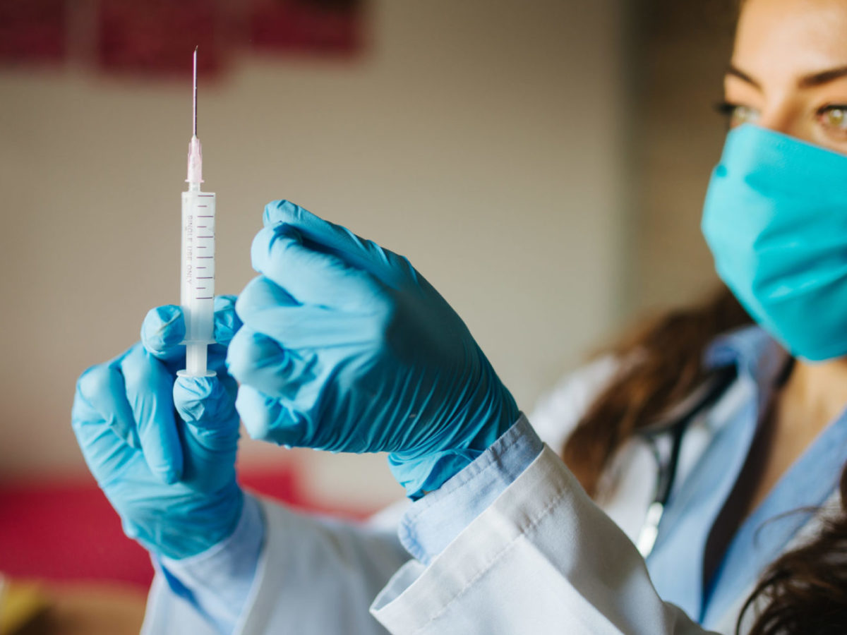 Health care worker, wearing a mask and gloves, holding a needle up.