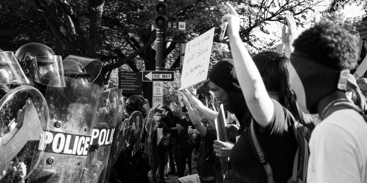 Photo of police with shields and batons stand across protestors holding up signs.