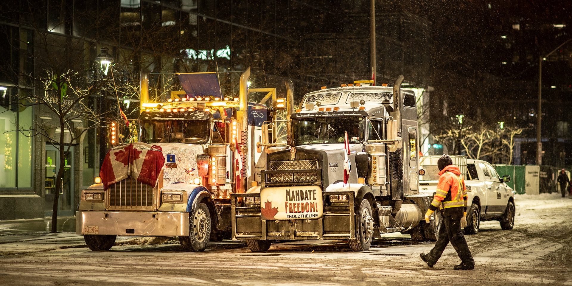 Photo of two trucks in the night time decorated with Canadian flags and protest posters. Man in reflective jackets walks nearby trucks.