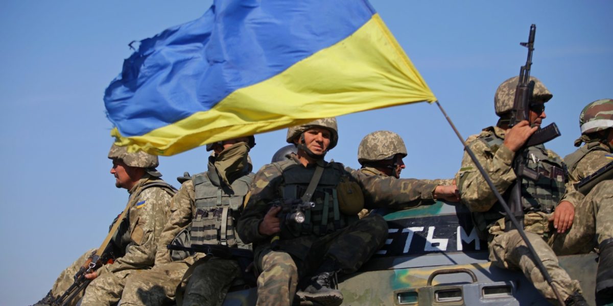 Photo of multiple soldiers sit on top of a tank holding weapons and large Ukrainian national flag.