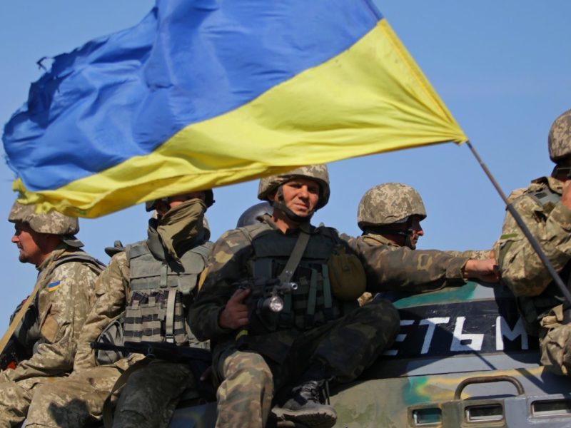 Photo of multiple soldiers sit on top of a tank holding weapons and large Ukrainian national flag.