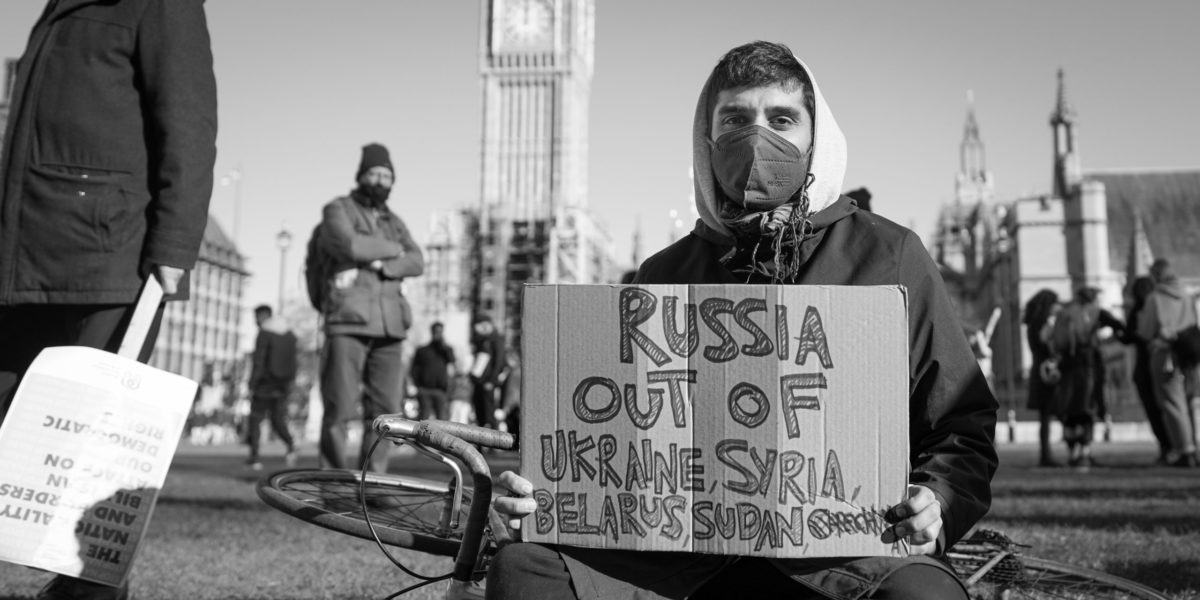 Photo of a man sitting on the grass, Big Ben behind him, holding anti-war poster.