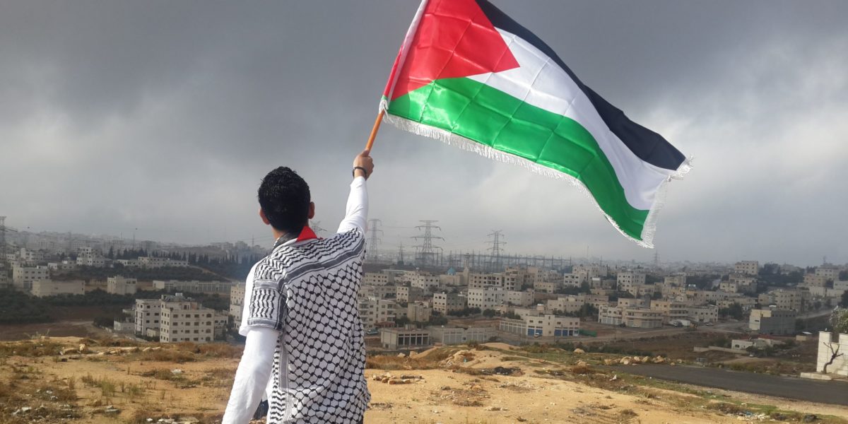 A man raises the Palestinian flag while a Palestinian city is in the background.