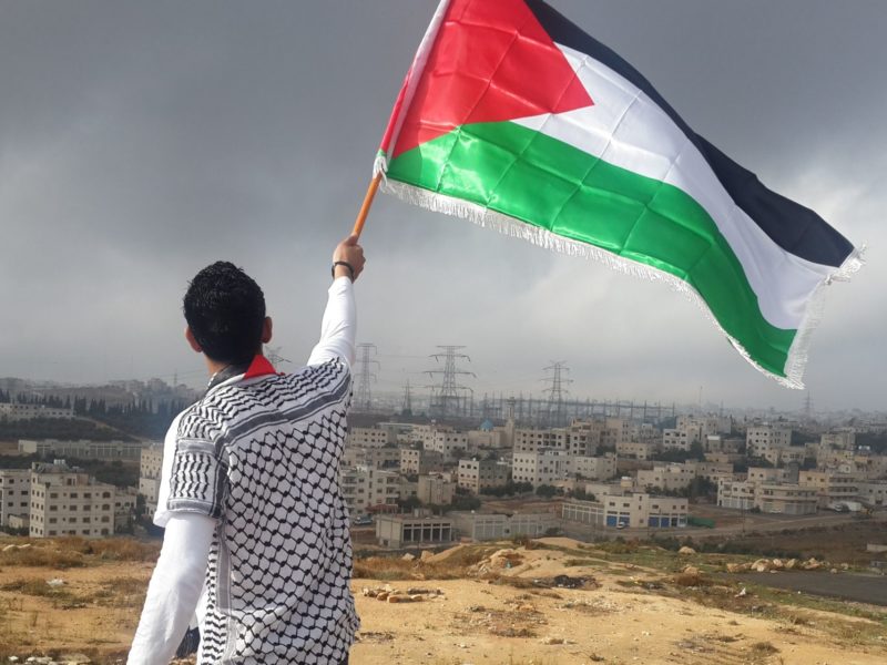 A man raises the Palestinian flag while a Palestinian city is in the background.