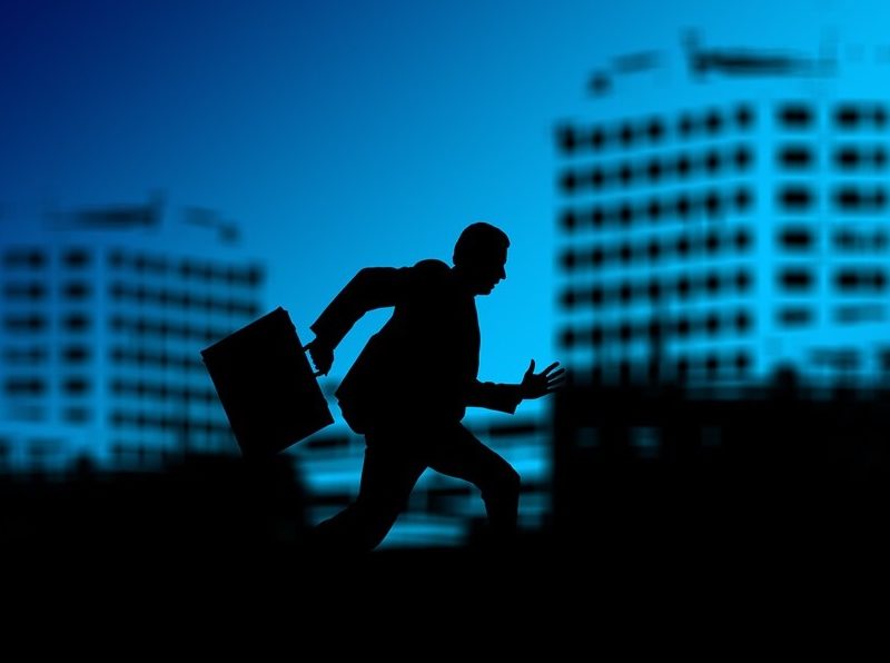 Image shows silhouette of person with briefcase, depicting a billionaire, in front of office buildings.