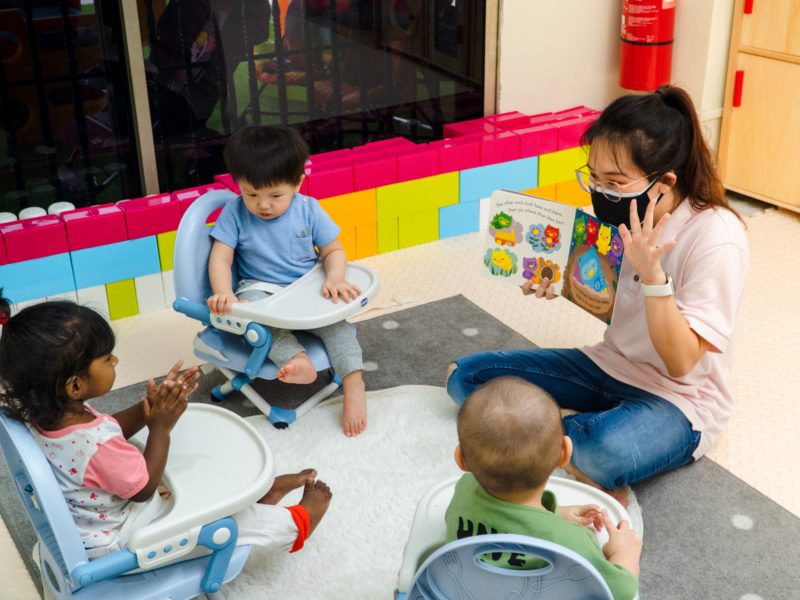 Photo of a woman wearing a mask and sitting on the floor showing a picture book to three young children.