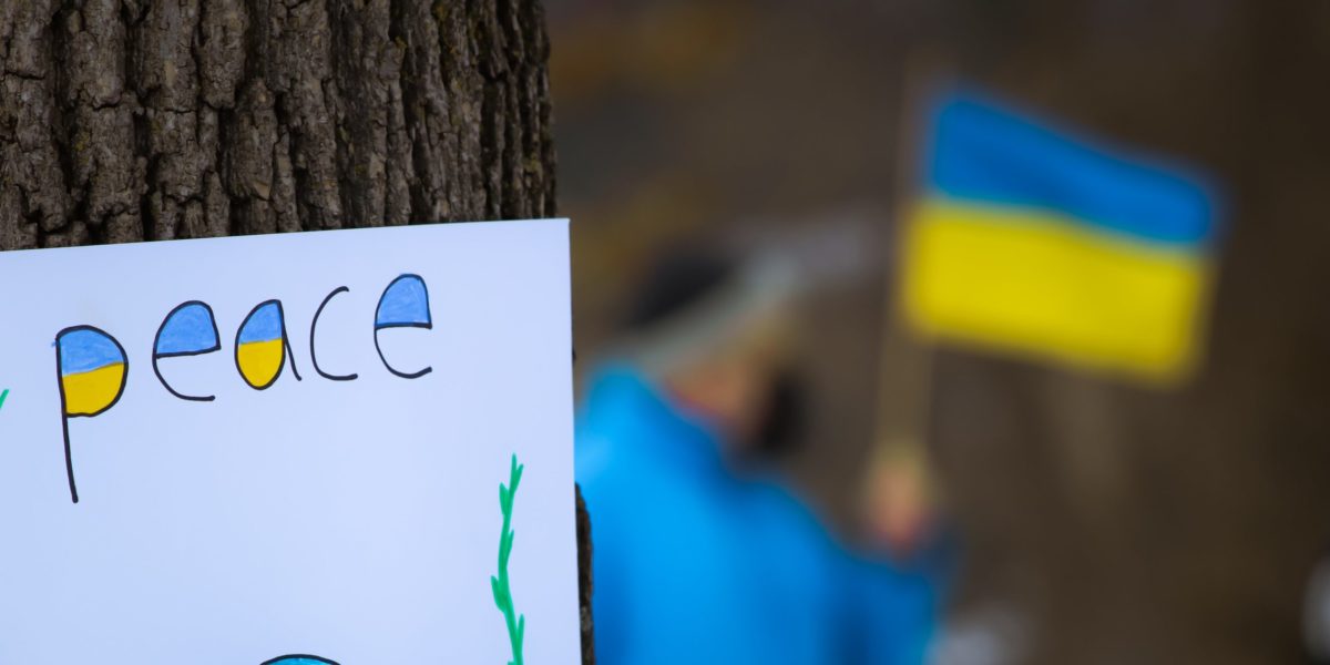 A hand-made "peace" sign zipped to a tree bark with a man holding the Ukrainian flag in the backdrop.