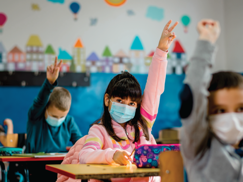 Photo of children in a classroom wearing medical masks and raising their hands.