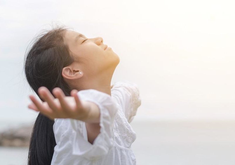 Photo of a woman standing in front of the ocean with her arms lifted up and her head tilted back like she is breathing clean air.