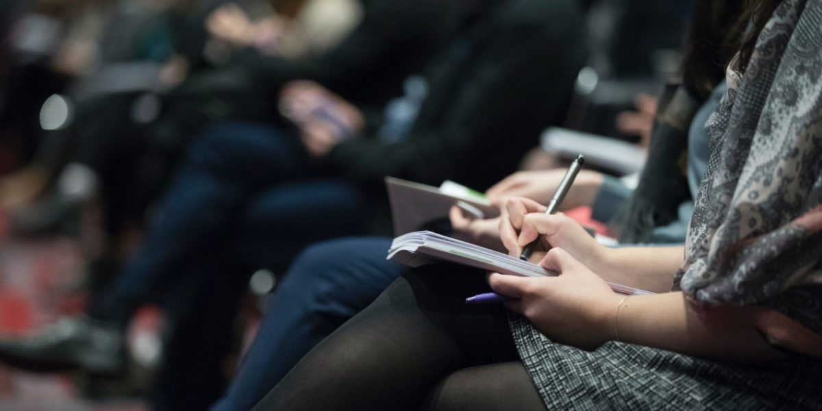 A photo of a crowd of people sitting together, writing on notepads.