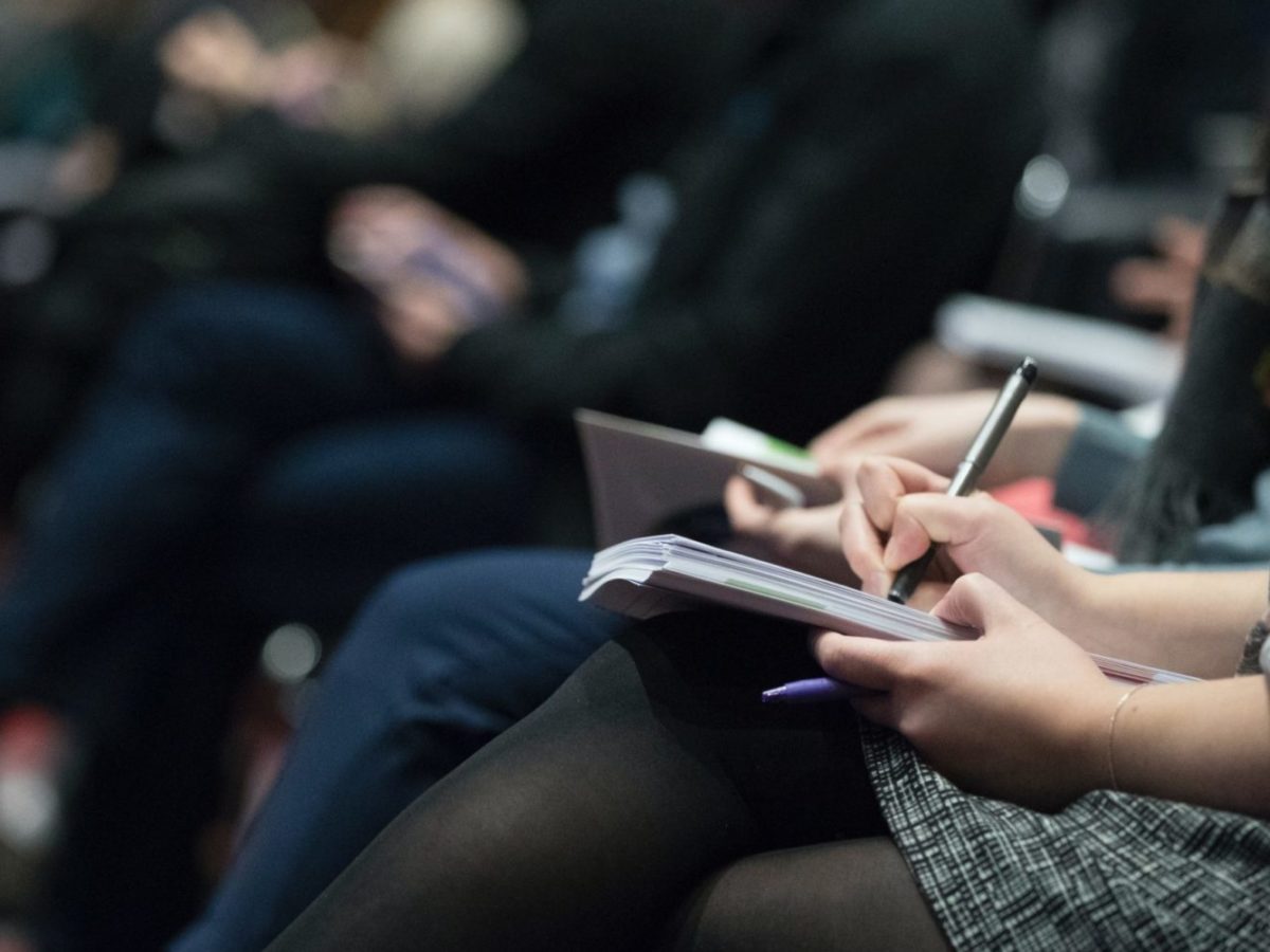 A photo of a crowd of people sitting together, writing on notepads.