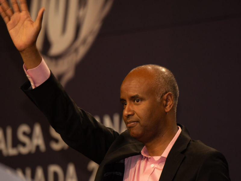 A photo of Ahmed Hussen standing at a podium waves hand.