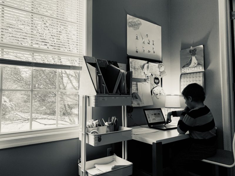 Dark photo of a child sitting at a desk at home pushing a button on a computer for virtual learning.