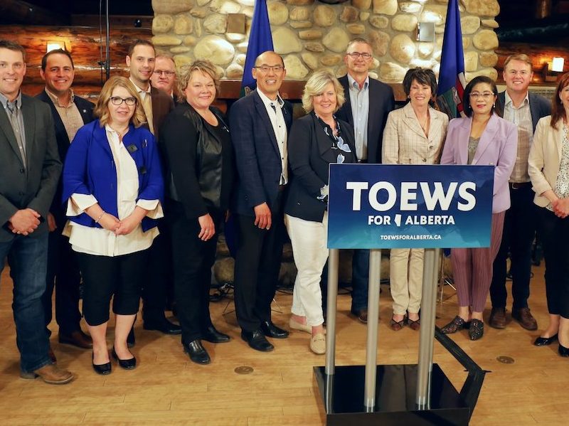 A photo of UCP leadership candidate Travis Toews, between the blue flags, with 14 of his supporters from the Government Caucus in the Alberta Legislature.