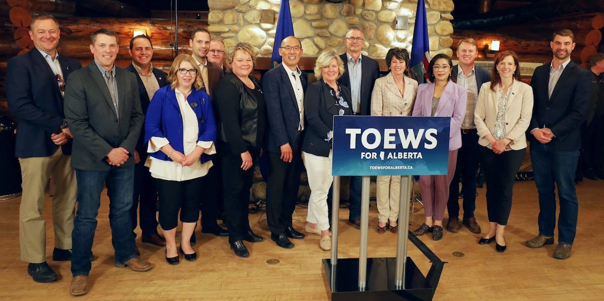 A photo of UCP leadership candidate Travis Toews, between the blue flags, with 14 of his supporters from the Government Caucus in the Alberta Legislature.