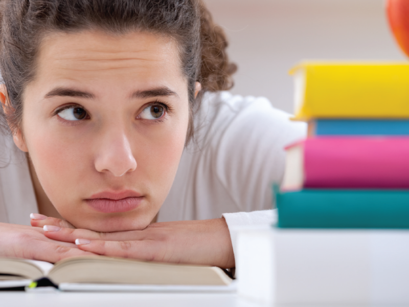 A young women rests her chin on her hands on an open book looking at a pile of books on her desk.