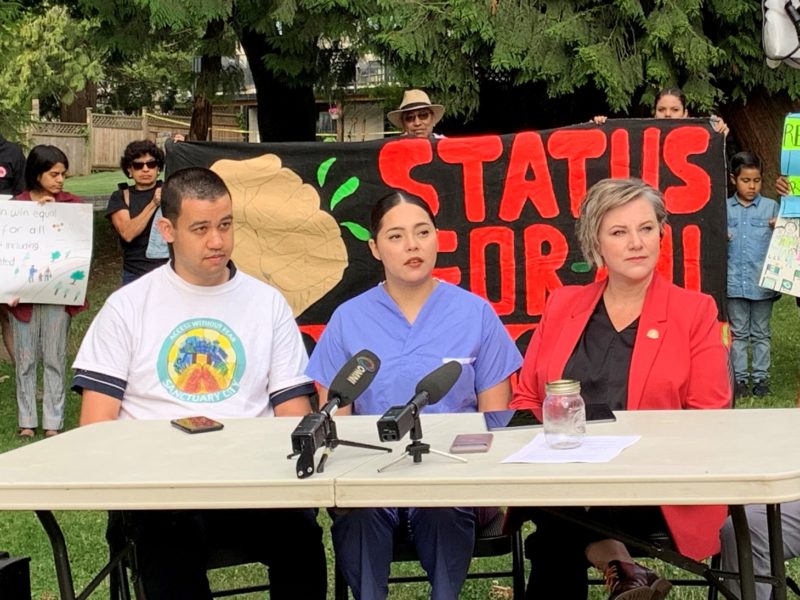 A man in a white T-shirt with the words "access without fear Sanctuary City", a woman wearing blue scrubs and a woman in a red blazer sit at a table with microphones on it outside with people holding a banner reading "status for all" and signs behind them.