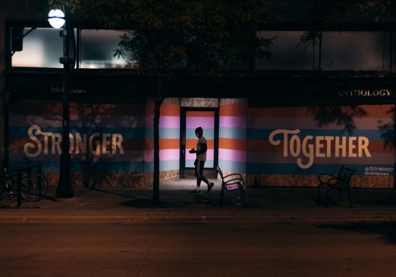 Night time photo of a person silhouetted in a doorway with blue, pink and red stripes behind them with the words stronger together written on the building on either side of the doorway
