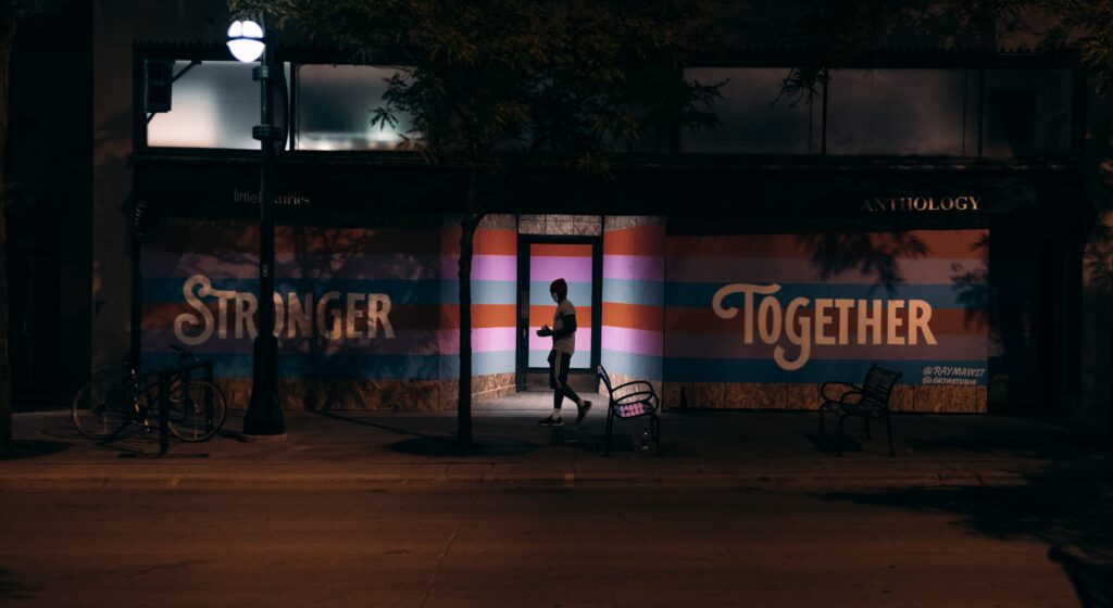 Night time photo of a person silhouetted in a doorway with blue, pink and red stripes behind them with the words stronger together written on the building on either side of the doorway