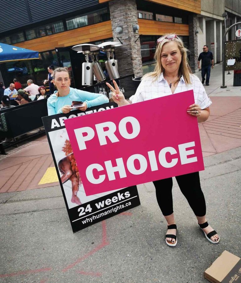 Image of a woman holding a sign reading "pro choice" and giving the peace sign of two fingers while the sign is blocking a graphic anti-abortion sign held by a person standing behind her.