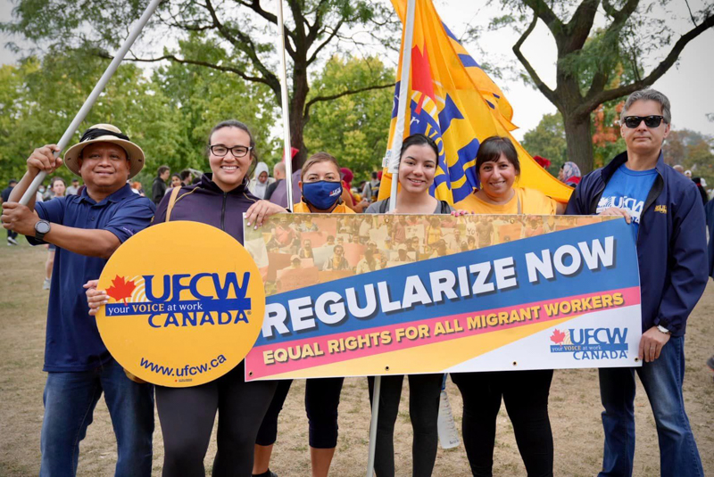 Photo of six migrant worker activists standing outside holding a UFCW Canada sign and flag as well as a banner reading regularization now equal rights for all migrant workers.