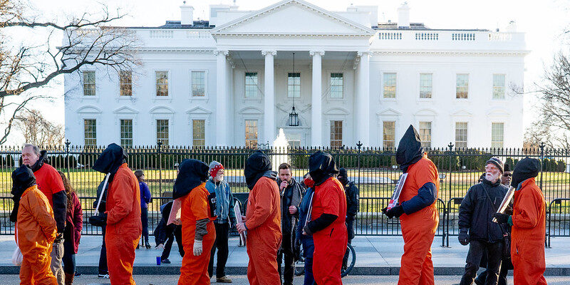 Seven figures in orange prison jumpsuits and black head coverings protest in front of the White House.