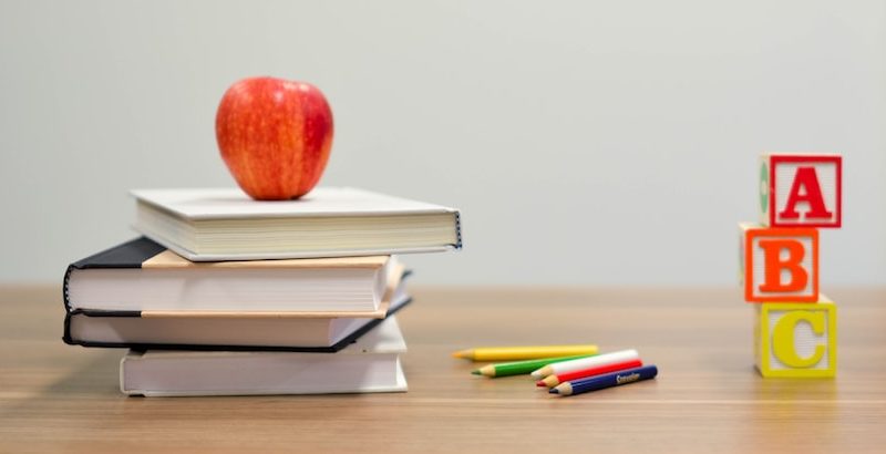 An image of a desk with a stack of books, an apple and building blocks that have the first three letters of the alphabet