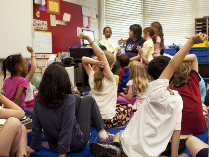 An image of elementary student sitting on a carpet listening to a teacher.