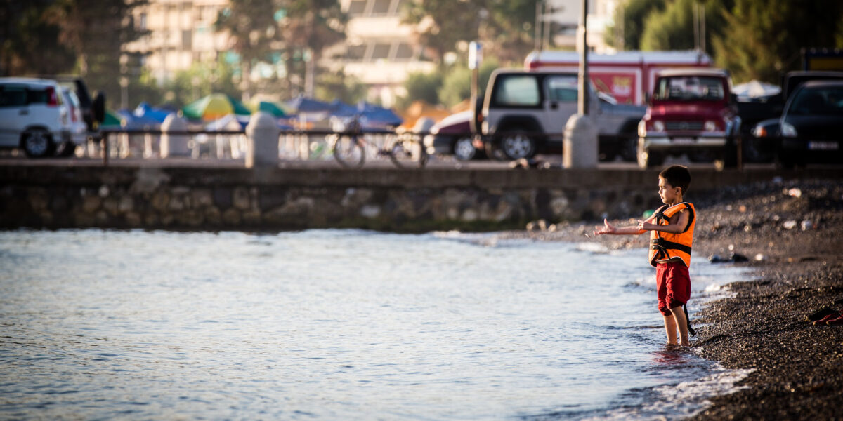 A photo of a boy standing on the shore of a beach in Kos, Greece. He faces back towards the Turkish coast across water that his family crossed on a migrant ship.