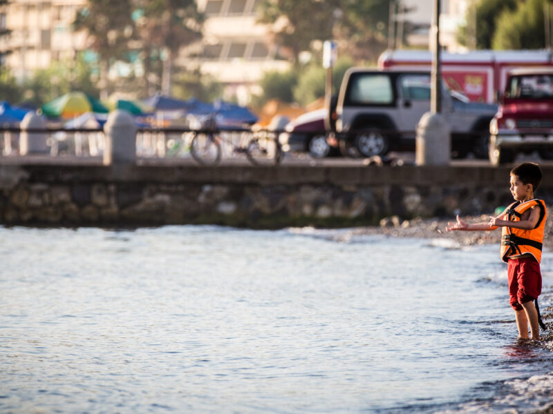 A photo of a boy standing on the shore of a beach in Kos, Greece. He faces back towards the Turkish coast across water that his family crossed on a migrant ship.