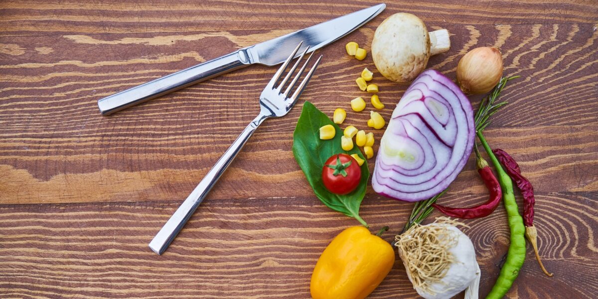 A knife and fork sit next to a selection of vegetables including an onion, yellow pepper, mushroom, and garlic, all part of a vegan diet.